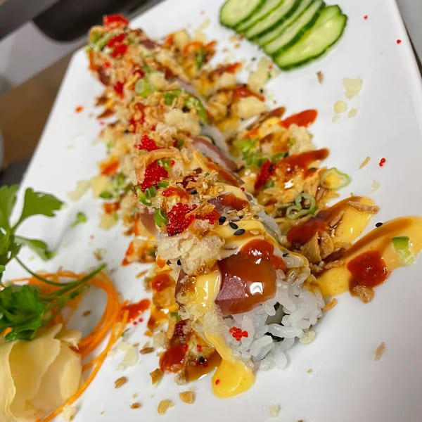 A long sushi roll with red roe, crispy toppings, and various sauces, garnished with cucumber and ginger.