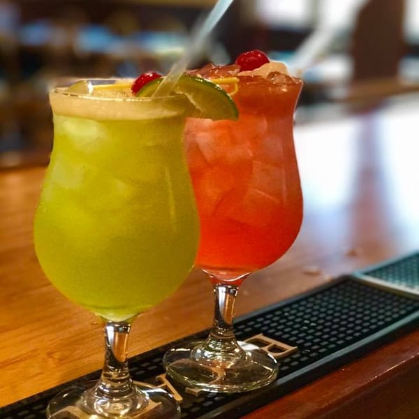 Two vibrant cocktails, one green and one red, garnished with lime and cherries on a bar.